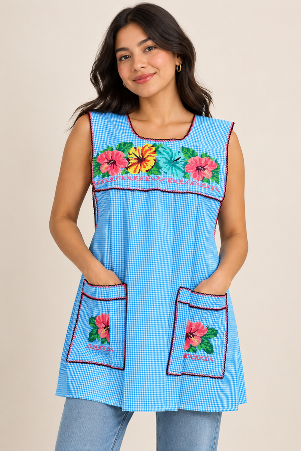 Woman wearing a handmade Mexican cross-stitch embroidered apron in baby blue gingham with colorful floral designs and front pockets.