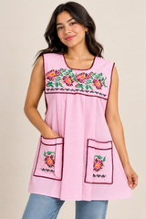 Woman wearing baby pink gingham Mexican apron with colorful floral cross-stitch embroidery and front pockets.