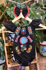 A black baby bodysuit with colorful floral embroidery, hanging on a wooden hanger with a matching white bow headband. The background features lush green houseplant leaves.