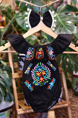 A black baby bodysuit with colorful floral embroidery, hanging on a wooden hanger with a matching white bow headband. The background features lush green houseplant leaves.