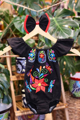 A black baby bodysuit with colorful floral embroidery, hanging on a wooden hanger with a matching white bow headband. The background features lush green houseplant leaves.