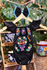 A black baby bodysuit with colorful floral embroidery, hanging on a wooden hanger with a matching white bow headband. The background features lush green houseplant leaves.