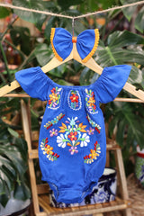 A blue baby bodysuit with colorful floral embroidery, hanging on a wooden hanger with a matching white bow headband. The background features lush green houseplant leaves.