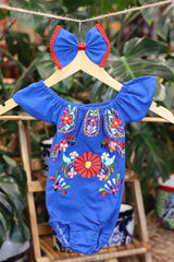 A blue baby bodysuit with colorful floral embroidery, hanging on a wooden hanger with a matching white bow headband. The background features lush green houseplant leaves.