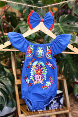 A blue baby bodysuit with colorful floral embroidery, hanging on a wooden hanger with a matching white bow headband. The background features lush green houseplant leaves.