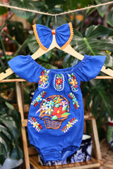 A blue baby bodysuit with colorful floral embroidery, hanging on a wooden hanger with a matching white bow headband. The background features lush green houseplant leaves.