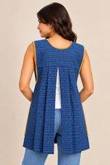 Back view of a blue plaid Mexican apron featuring an open-back design, pleated fabric, and colorful embroidered trim, worn over a white top and jeans.