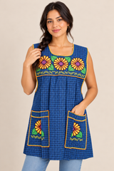 Model wearing a blue plaid Mexican apron with colorful yellow floral cross-stitch embroidery, two front pockets, and decorative trim.