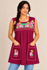 Model wearing a handmade burgundy plaid Mexican apron with colorful floral embroidery, hands resting inside the front pockets.