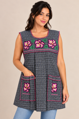 Model posing in a gray plaid Mexican apron with colorful floral embroidery, two front pockets, and blue jeans.