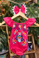 A hot pink baby bodysuit with colorful floral embroidery, hanging on a wooden hanger with a matching white bow headband. The background features lush green houseplant leaves.