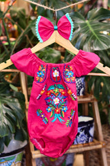 A hot pink baby bodysuit with colorful floral embroidery, hanging on a wooden hanger with a matching white bow headband. The background features lush green houseplant leaves.