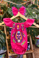 A hot pink baby bodysuit with colorful floral embroidery, hanging on a wooden hanger with a matching white bow headband. The background features lush green houseplant leaves.