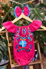 A hot pink baby bodysuit with colorful floral embroidery, hanging on a wooden hanger with a matching white bow headband. The background features lush green houseplant leaves.
