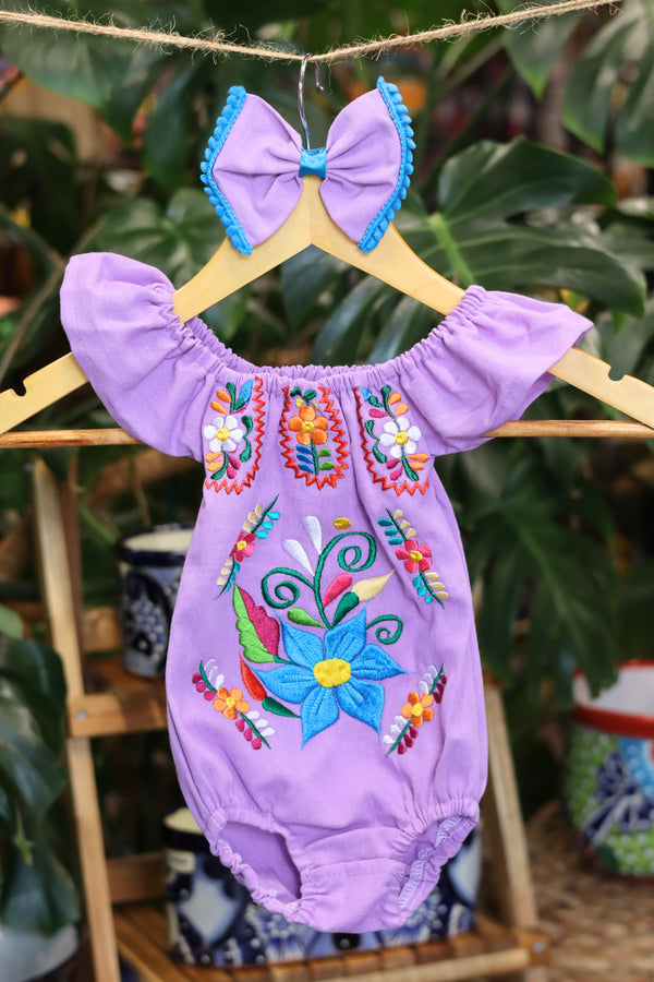A lavender baby bodysuit with colorful floral embroidery, hanging on a wooden hanger with a matching white bow headband. The background features lush green houseplant leaves.