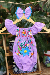 A lavender baby bodysuit with colorful floral embroidery, hanging on a wooden hanger with a matching white bow headband. The background features lush green houseplant leaves.