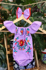 A lavender baby bodysuit with colorful floral embroidery, hanging on a wooden hanger with a matching white bow headband. The background features lush green houseplant leaves.