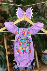 A lavender baby bodysuit with colorful floral embroidery, hanging on a wooden hanger with a matching white bow headband. The background features lush green houseplant leaves.