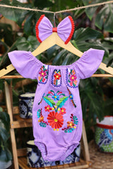 A lavender baby bodysuit with colorful floral embroidery, hanging on a wooden hanger with a matching white bow headband. The background features lush green houseplant leaves.