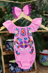 A pink baby bodysuit with colorful floral embroidery, hanging on a wooden hanger with a matching white bow headband. The background features lush green houseplant leaves.