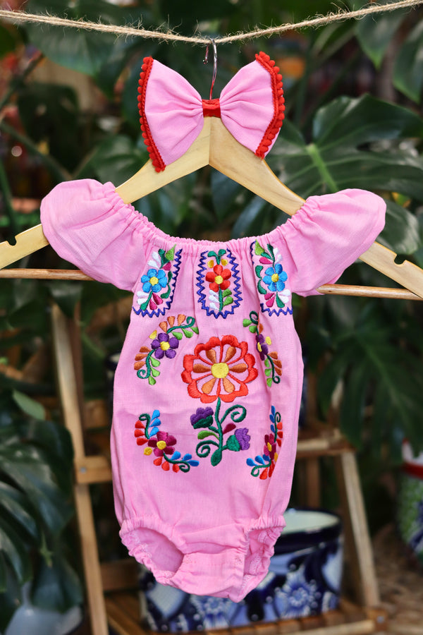 A pink baby bodysuit with colorful floral embroidery, hanging on a wooden hanger with a matching white bow headband. The background features lush green houseplant leaves.