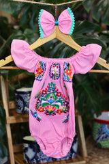 A pink baby bodysuit with colorful floral embroidery, hanging on a wooden hanger with a matching white bow headband. The background features lush green houseplant leaves.