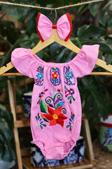 A pink baby bodysuit with colorful floral embroidery, hanging on a wooden hanger with a matching white bow headband. The background features lush green houseplant leaves.