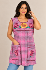 Model wearing a pink plaid Mexican apron with colorful floral cross-stitch embroidery, two front pockets, and decorative trim.