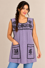 Model wearing a purple gingham Mexican apron with black floral cross-stitch embroidery, two front pockets, and a side braid.