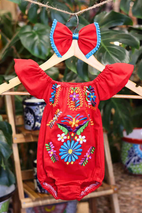 A red baby bodysuit with colorful floral embroidery, hanging on a wooden hanger with a matching white bow headband. The background features lush green houseplant leaves.