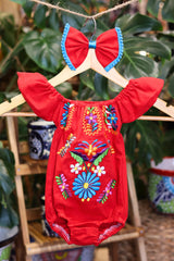 A red baby bodysuit with colorful floral embroidery, hanging on a wooden hanger with a matching white bow headband. The background features lush green houseplant leaves.