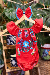 A red baby bodysuit with colorful floral embroidery, hanging on a wooden hanger with a matching white bow headband. The background features lush green houseplant leaves.