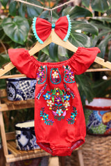A red baby bodysuit with colorful floral embroidery, hanging on a wooden hanger with a matching white bow headband. The background features lush green houseplant leaves.