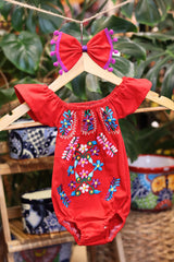 A red baby bodysuit with colorful floral embroidery, hanging on a wooden hanger with a matching white bow headband. The background features lush green houseplant leaves.