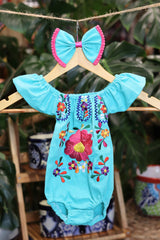 A turquoise baby bodysuit with colorful floral embroidery, hanging on a wooden hanger with a matching white bow headband. The background features lush green houseplant leaves.