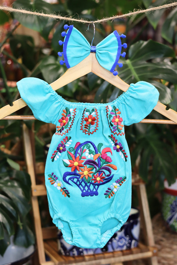 A turquoise baby bodysuit with colorful floral embroidery, hanging on a wooden hanger with a matching white bow headband. The background features lush green houseplant leaves.