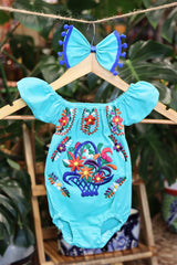 A turquoise baby bodysuit with colorful floral embroidery, hanging on a wooden hanger with a matching white bow headband. The background features lush green houseplant leaves.