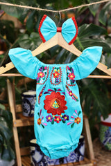 A turquoise baby bodysuit with colorful floral embroidery, hanging on a wooden hanger with a matching white bow headband. The background features lush green houseplant leaves.