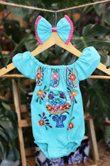 A turquoise baby bodysuit with colorful floral embroidery, hanging on a wooden hanger with a matching white bow headband. The background features lush green houseplant leaves.