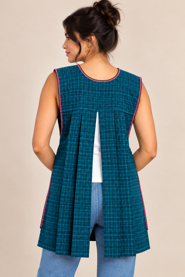 Back view of a dark teal plaid Mexican apron featuring a sleeveless design, open pleated back, and decorative trim, modeled with blue jeans.