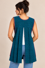 Back view of a dark teal plaid Mexican apron featuring a sleeveless design, open pleated back, and decorative trim, modeled with blue jeans.