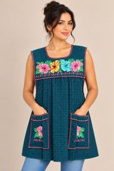 Model wearing a dark teal plaid Mexican apron with colorful floral cross-stitch embroidery, pink trim, and two front pockets.