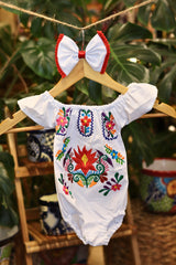 A white baby bodysuit with colorful floral embroidery, hanging on a wooden hanger with a matching white bow headband. The background features lush green houseplant leaves.