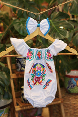 A white baby bodysuit with colorful floral embroidery, hanging on a wooden hanger with a matching white bow headband. The background features lush green houseplant leaves.