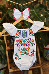 A white baby bodysuit with colorful floral embroidery, hanging on a wooden hanger with a matching white bow headband. The background features lush green houseplant leaves.
