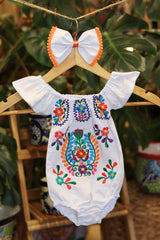 A white baby bodysuit with colorful floral embroidery, hanging on a wooden hanger with a matching white bow headband. The background features lush green houseplant leaves.