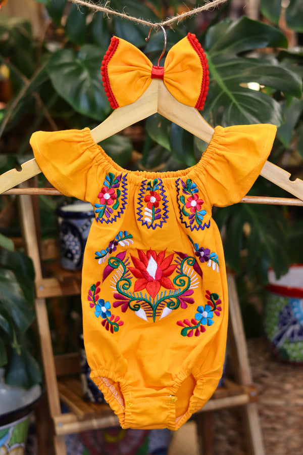 A yellow baby bodysuit with colorful floral embroidery, hanging on a wooden hanger with a matching white bow headband. The background features lush green houseplant leaves.