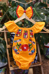 A yellow baby bodysuit with colorful floral embroidery, hanging on a wooden hanger with a matching white bow headband. The background features lush green houseplant leaves.