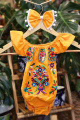 A yellow baby bodysuit with colorful floral embroidery, hanging on a wooden hanger with a matching white bow headband. The background features lush green houseplant leaves.