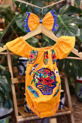A yellow baby bodysuit with colorful floral embroidery, hanging on a wooden hanger with a matching white bow headband. The background features lush green houseplant leaves.